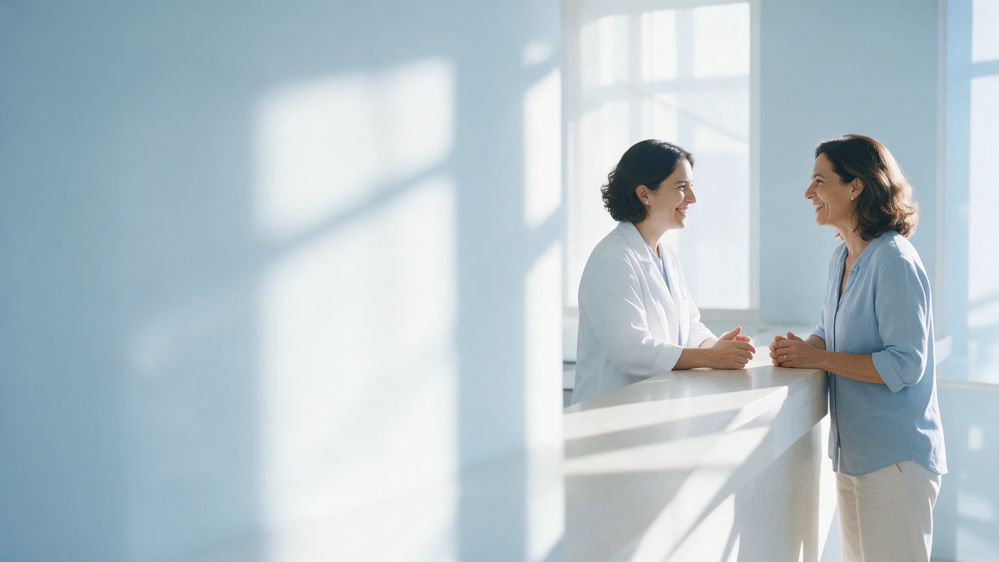 Farmacêutica de jaleco e cliente conversam sorrindo em um balcão de pedra clara, sob a luz da manhã